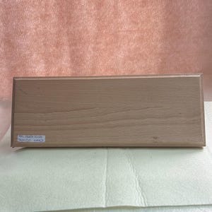 May include: A light brown wooden plaque, rectangular in shape, resting on a stack of pale yellow fabric. The plaque has a small label and is set against a backdrop of pink paper.