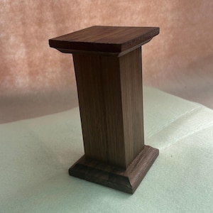 May include: A small, dark brown wooden pedestal with a square top and base. The pedestal has a rectangular column and is likely used for displaying small items. The wood grain is visible.