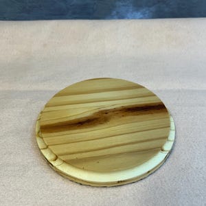 May include: A circular wooden coaster with a light brown finish and a darker wood grain pattern. The coaster has a decorative edge and is made of natural wood.