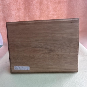 May include: A rectangular wooden box with a light brown finish. The box has a label that reads "XL PLINTH 205x150 OAK". The wood grain is visible on the surface. The box is sitting on a light green surface.