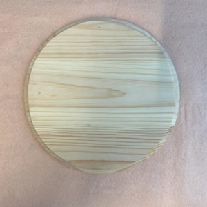 May include: A circular wooden serving board with a clear, rounded edge. The wood grain is visible, with light brown and tan tones. The board is on a pink surface.