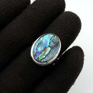 May include: An oval-shaped ring featuring a polished abalone shell inlay with iridescent swirls of blue, green, and purple. The shell is set within a smooth, silver-toned metal bezel, creating a reflective surface that highlights the natural patterns.