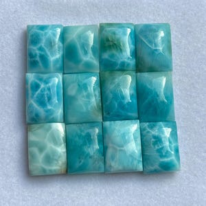May include: A selection of twelve rectangular larimar gemstones. The stones exhibit varying shades of blue and turquoise with white veining, producing a marbled appearance. The polished surfaces reflect light, highlighting the natural patterns within the stones.