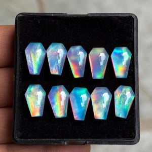 May include: Ten coffin-shaped iridescent gemstones arranged in two rows of five inside a black display box. Each stone features a smooth, polished surface with shifting blue, green, and pink light reflections that create a vibrant, glowing effect.