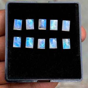 Könnte beinhalten: Zehn rechteckige Mondstein-Edelsteine, die jeweils ein faszinierendes blau-weißes Schimmern aufweisen. Die Steine sind in einem schwarzen Display-Etui angeordnet, das ihre polierten Oberflächen und ihre natürliche Schönheit zur Geltung bringt. Die Steine sind rechteckig.