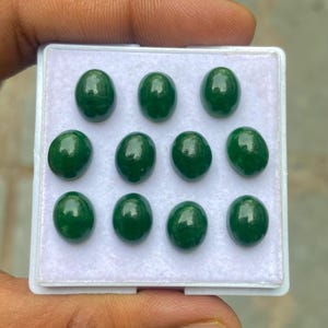 Puede incluir: Una colección de once gemas ovaladas de color verde oscuro, presentadas en una bandeja blanca. Las piedras pulidas tienen una superficie lisa y brillante, que refleja la luz. Las gemas están dispuestas en tres filas.