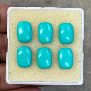 May include: Six polished, turquoise gemstones with a rectangular shape and rounded corners are displayed in a white plastic container with a beige foam insert. The gemstones have a glossy finish and are evenly spaced.