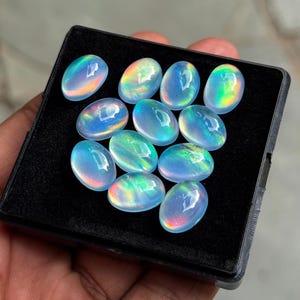 May include: A collection of oval-shaped opals displayed in a black tray. The opals exhibit a play of color, with iridescent flashes of blue, green, and yellow. The gemstones are smooth and polished, reflecting light.