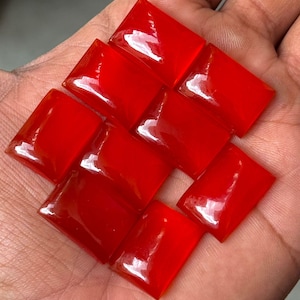 May include: Ten glossy, bright red rectangular gemstones are arranged in a woven pattern. The polished stones reflect light, creating a shimmering effect. The arrangement is held in a hand, highlighting the colour and shape of the stones.