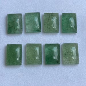 May include: Eight green aventurine gemstone cabochons, rectangular in shape, are arranged on a white surface. The stones vary in shade from light to dark green, with a slightly translucent appearance. These polished stones are ideal for jewelry making.