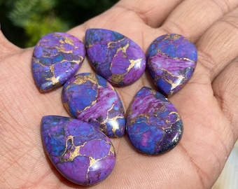 May include: Six teardrop-shaped gemstones with a vibrant mix of purple, blue, and gold hues. The stones have a polished surface, showcasing the natural patterns and colors. The gemstones are arranged on a hand, with a blurred green background.