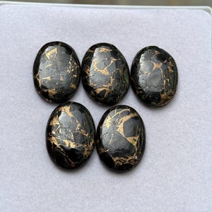 May include: Five oval black stones with gold veining. The polished stones have a glossy finish. The gold veins create a striking contrast against the dark background. Suitable for jewellery making or other craft projects.