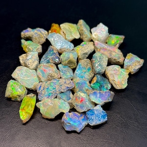 May include: A collection of raw, unpolished opal stones featuring a variety of irregular shapes and sizes. Each stone displays a translucent to opaque base with vibrant flashes of iridescent color, including green, blue, orange, and yellow.