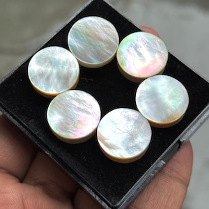 Könnte beinhalten: Sechs runde, irisierende Cabochons aus Perlmutt in einer schwarzen Schale. Die Cabochons haben einen perlmuttartigen Glanz mit rosa, grünen und gelben Farbtönen. Jeder Cabochon hat einen Durchmesser von etwa 1,9 cm.