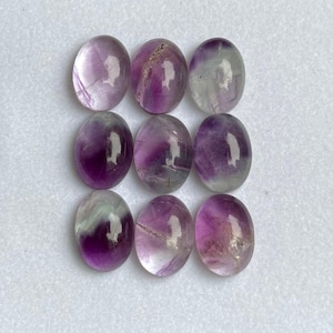 May include: Nine oval-shaped fluorite gemstones. The stones display a mix of purple, lavender, and clear hues, with natural veining patterns. The polished stones are arranged in a three-by-three grid, showcasing their translucent quality.