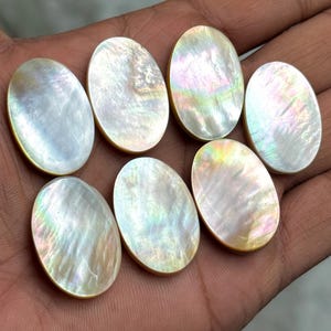 Könnte beinhalten: Sieben ovale Cabochons aus Perlmutt. Die schillernden Edelsteine zeigen eine Reihe von Farben, darunter Weiß, Gold und Hauch von Grün und Rosa. Die Cabochons sind glatt und poliert, mit einem dezenten Glanz.