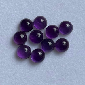 Könnte beinhalten: Eine Sammlung von zehn polierten, runden Amethyst-Edelsteinen. Die tiefvioletten Steine haben eine glänzende Oberfläche und sind auf einer hellgrauen Oberfläche angeordnet. Jeder Stein hat eine glatte, gewölbte Form.