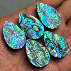 May include: Six iridescent abalone shell teardrop cabochons. The polished stones display a swirling pattern of blues, greens, and purples. The cabochons are approximately 1.5 inches long.