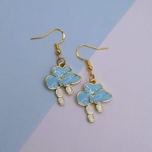 May include: A pair of gold-coloured hook earrings. Each earring features a light blue cloud design with white raindrop accents. The earrings have a shiny, metallic finish and are set against a light blue and white background.