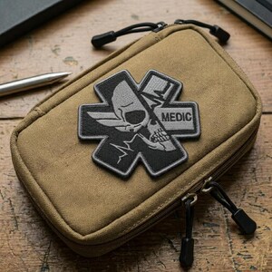 May include: Tan zippered pouch with a black and gray patch featuring a skull within a cross design, with the word "MEDIC". The pouch has a black zipper pull and is likely for medical supplies.
