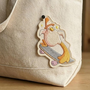 May include: Embroidered patch featuring a penguin riding a skateboard. The penguin is wearing a traffic cone hat and has pink, yellow, and white details. The skateboard is gray with pink wheels. The patch is attached to a beige tote bag.