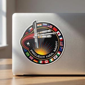 May include: A circular sticker featuring a space-themed design with the text "PROJECT HAIL MARY." The design includes a rocket, a rising sun, and flags of various countries. The sticker is on a laptop.