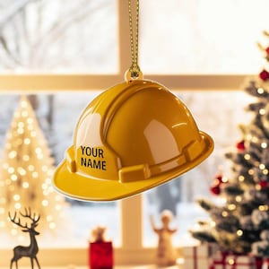 May include: A yellow construction helmet ornament with the text "YOUR NAME" in black. The ornament hangs from a gold string. The background includes a Christmas tree and a window.