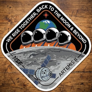 May include: A diamond-shaped sticker featuring four astronauts in orange suits and white helmets, with the Earth and a star in the background. The sticker reads "WE RISE TOGETHER, BACK TO THE MOON & BEYOND" and "ARTEMIS II". A spacecraft is also depicted.