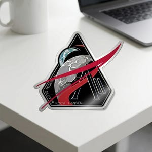 May include: A sticker featuring a triangular design with a black background and a stylized image of the moon and Earth. Red streaks cross the design, and the names GLOVER, KOCH, HANSEN, and WISEMAN are printed on the sticker.