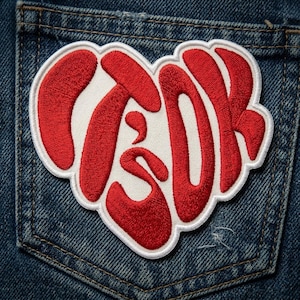 May include: Embroidered heart-shaped patch with the word "TSOK" in red, stylized lettering. The patch has a white border and is set against a blue denim background, likely a pocket. The patch is a decorative item.