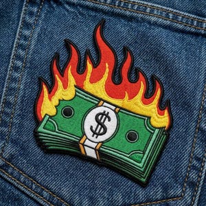 May include: Embroidered patch featuring a stack of green dollar bills with a white band and a dollar sign, engulfed in flames. The flames are red and yellow. The patch is on a blue denim background.