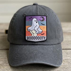 May include: Gray baseball cap with a patch featuring a cartoon ghost with the text "BOOOOOOTY". The patch has a colorful background with a moon, stars, and bats. The cap is made of a soft, durable material.