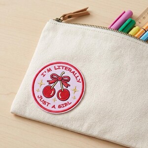 May include: A cream-colored canvas pencil case with a zipper and a leather pull tab. A circular patch with the text "I'M LITERALLY JUST A GIRL" and a cherry design is attached to the case. Several pens and a ruler are visible inside.