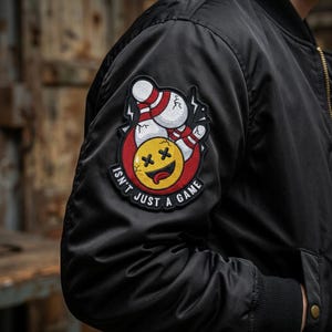 May include: Black bomber jacket with a bowling-themed patch. The patch features a smiling yellow face with crossed-out eyes, bowling pins, and the text "Isn't Just A Game" in white.