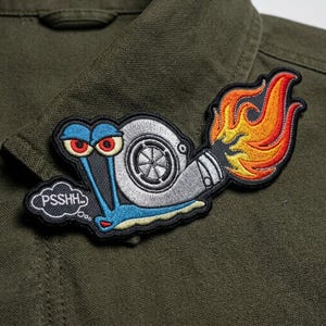 May include: Embroidered patch featuring a cartoon snail with a turbocharger and flames. The snail is blue with red eyes, a silver turbo, and a flame design in orange, yellow, and red. A speech bubble says "PSSHH."