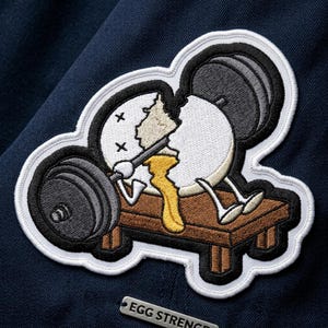 May include: Embroidered patch featuring a cartoon egg lifting weights on a bench press. The egg is cracked, with yolk dripping out. The patch has a white border and the text "EGG STRENGTH" on a small metal tag.