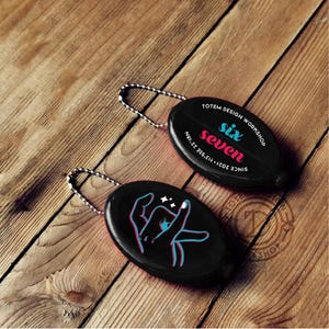 Puede incluir: Dos llaveros ovalados negros con cadenas plateadas sobre una superficie de madera. Un llavero presenta el texto "six seven" y "TOTEM DESIGN WORKSHOP". El otro tiene un gráfico de mano con estrellas.