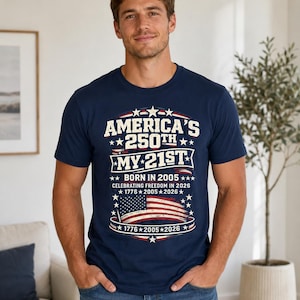 Könnte beinhalten: Ein kurzärmeliges T-Shirt in Marineblau mit einem Grafikdruck, der den Text AMERICA'S 250TH MY 21ST BORN IN 2005 CELEBRATING FREEDOM IN 2026 1776 2005 2026 sowie eine abgenutzte amerikanische Flagge, Sterne und dekorative Banner zeigt.