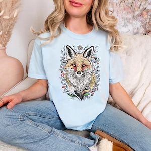 Può includere: T-shirt azzurra con una dettagliata illustrazione di una volpe circondata da elementi floreali. La volpe ha un mantello marrone-rossiccio, orecchie nere e petto bianco. Il disegno è incorniciato da foglie verdi e piccoli fiori.