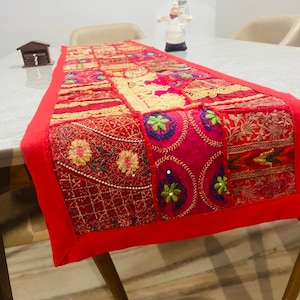 Op de afbeelding: Een kleurrijke patchwork tafelloper in rood en diverse kleuren. De loper heeft gedetailleerde borduursels en patronen in rood, goud en roze. Het heeft een effen rode rand. De loper is ongeveer 152 cm lang.