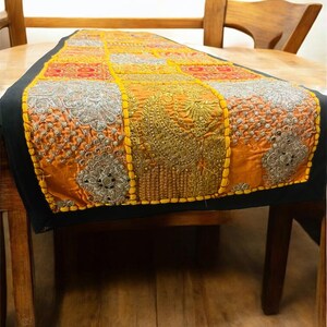 Könnte beinhalten: Ein orangefarbener und goldfarbener Tischläufer mit aufwendigen Stickmustern und einem schwarzen Rand. Der Läufer hat ein Patchwork-Design mit silbernen und goldenen Akzenten, das jedem Esstisch einen Hauch von Eleganz verleiht. Der Läufer liegt auf einem Holztisch.