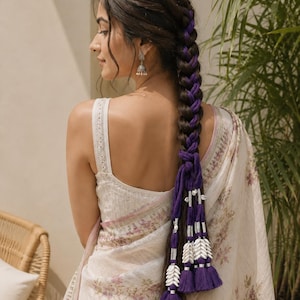 Può includere: Una treccia lunga e spessa intrecciata con un cordoncino di tessuto viola intenso, caratterizzata da accenti in metallo argentato, decorazioni con conchiglie bianche e diverse nappe in tessuto viola appese all'estremità della treccia.