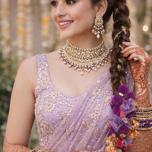 Può includere: Una blusa senza maniche e una gonna abbinata color lavanda con intricati ricami dorati e paillettes. L'insieme è completato da una collana girocollo dorata, orecchini pendenti abbinati, un maang tikka e braccialetti impilati viola e oro.