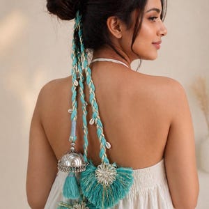 Può includere: Un accessorio per capelli composto da tre fili intrecciati di cordoncino metallico turchese e oro, decorato con piccole conchiglie bianche. Le trecce terminano con due grandi nappe turchesi con tappi floreali color argento.