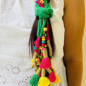 Può includere: Un accessorio per capelli composto da una spessa corda intrecciata verde annodata, decorata con numerose nappe colorate rosa, gialle e verdi, intervallate da tubicini metallici dorati, piccoli campanelli e varie perline multicolori.