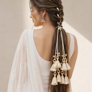 Può includere: Un'acconciatura lunga intrecciata decorata con un accessorio per capelli color crema caratterizzato da molteplici nappe pendenti, piccoli anelli di legno e dettagli metallici. L'accessorio è intrecciato nella treccia e scende lungo i capelli.