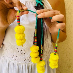 Può includere: Un accessorio per capelli con cordini intrecciati color ottanio inseriti nei capelli scuri, arricchito da spirali metalliche dorate e piccole perline di legno multicolori. Ogni cordino termina con un gruppo di tre pon pon giallo brillante.