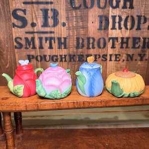 May include: Four ceramic flower-shaped teapots on a wooden shelf. The teapots are red, pink, blue, and yellow, with green detailing. The background features the text "S.B. Cough Drops Smith Brothers Poughkeepsie, N.Y."