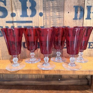 Puede incluir: Conjunto de seis copas de vino tinto rubí. Cada copa presenta un cuenco acampanado y acanalado, un tallo transparente y adornado y una base circular. Las copas están dispuestas en fila, mostrando su elegante diseño y color.