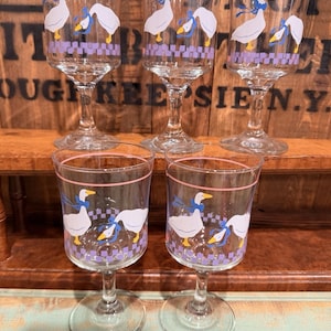 Op de afbeelding: Set van zes heldere wijnglazen. Elk glas heeft een wit ganzenontwerp met een blauwe strik en gele snavel. De glazen hebben een roze rand en een paars geruit patroon. Gepresenteerd op een houten plank.